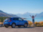 A person stands with arms raised next to a blue SUV, overlooking a scenic landscape of Aoraki Mount Cook and a turquoise lake