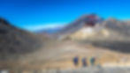 Three hikers traverse the barren trail of Ruapehu, framed by volcanic mountains under a clear blue sky.