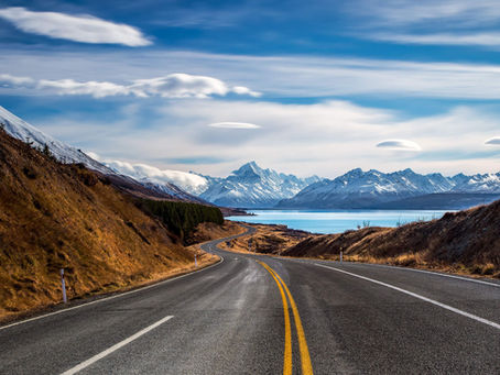 Open road leads to snow-capped mountains beside a blue lake under a sky with wispy clouds. Rocky hills flank the road, creating a serene scene.