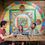 Thumbnail: Three people sit at an outdoor wooden table, smiling and chatting. Behind them, a colorful mosaic wall art adorns the bricks.