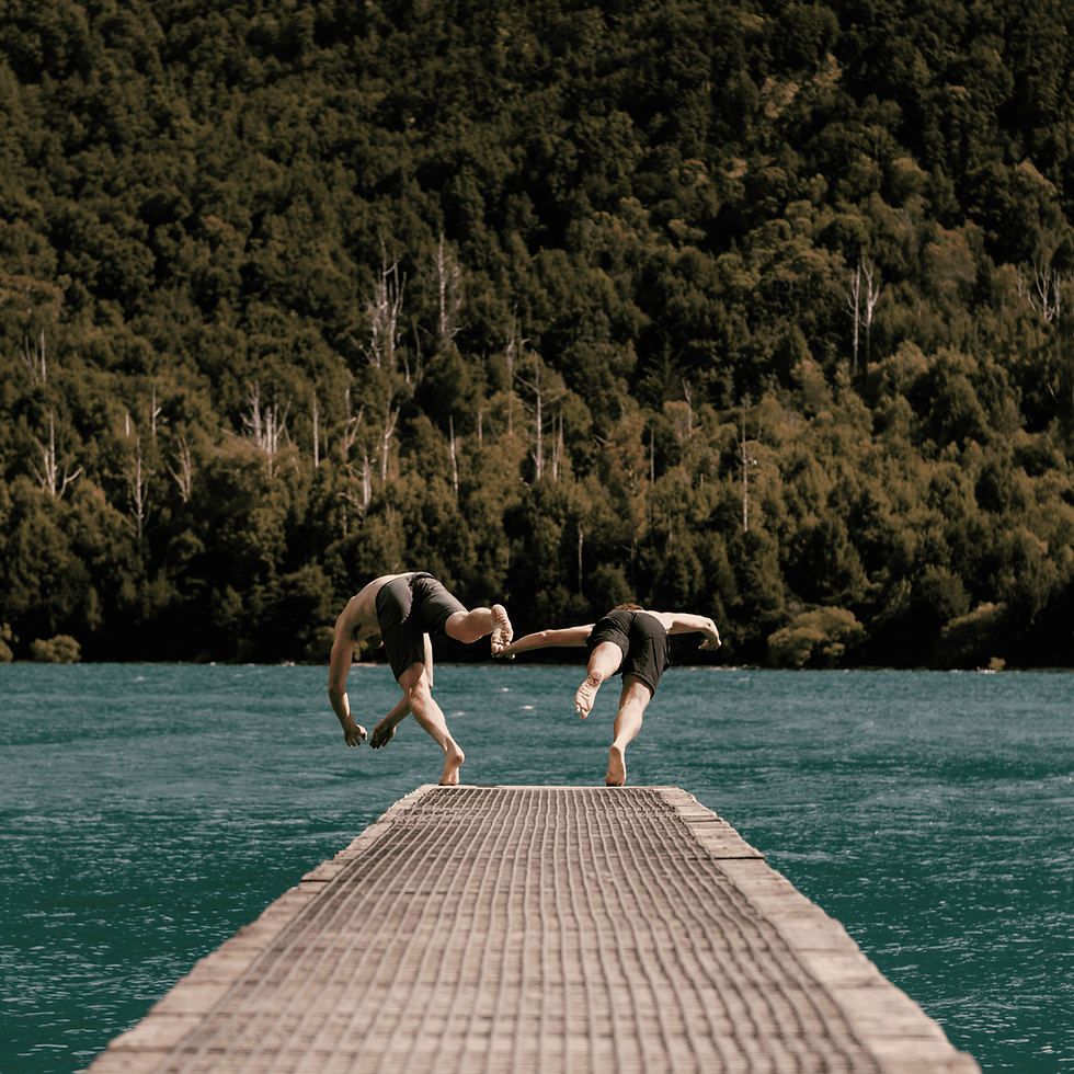 Two people dive off a wooden pier into a turquoise lake, surrounded by dense green forest. The scene is lively and adventurous.