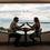 Thumbnail: A couple sits at a table by a large window overlooking the serene expanse of an ocean. Clinking their glasses together.n
