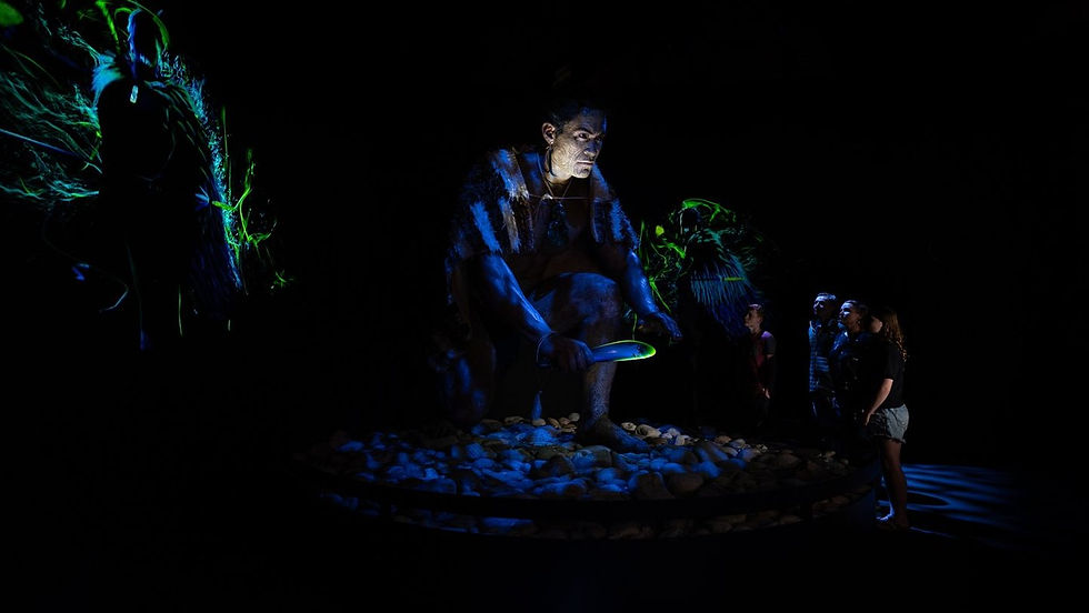 Thumbnail: In a dimly lit exhibit of Pounamu Pathway Mawhera, a large statue of a crouching figure holds a glowing object .