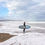 Thumbnail: A person stands with a blue surfboard, facing the ocean on a pebbled beach in Invercargill. Waves crash at their feet.