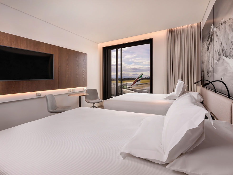Thumbnail: The Airport Overnighter room boasts two neatly made beds, a large wall-mounted TV, and a small round table with two chairs.