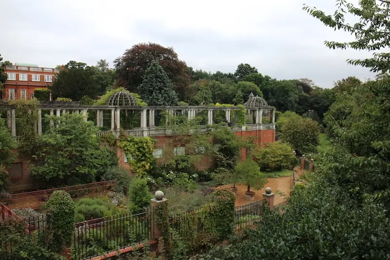 Lush garden with pergola and ivy-covered walls, surrounded by trees. Red brick building in the background under a cloudy sky.