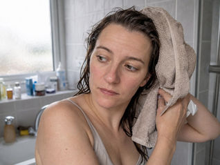 Woman drying wet hair with a beige towel in a bathroom. Background shows toiletries on a counter and frosted glass window. Calm mood.