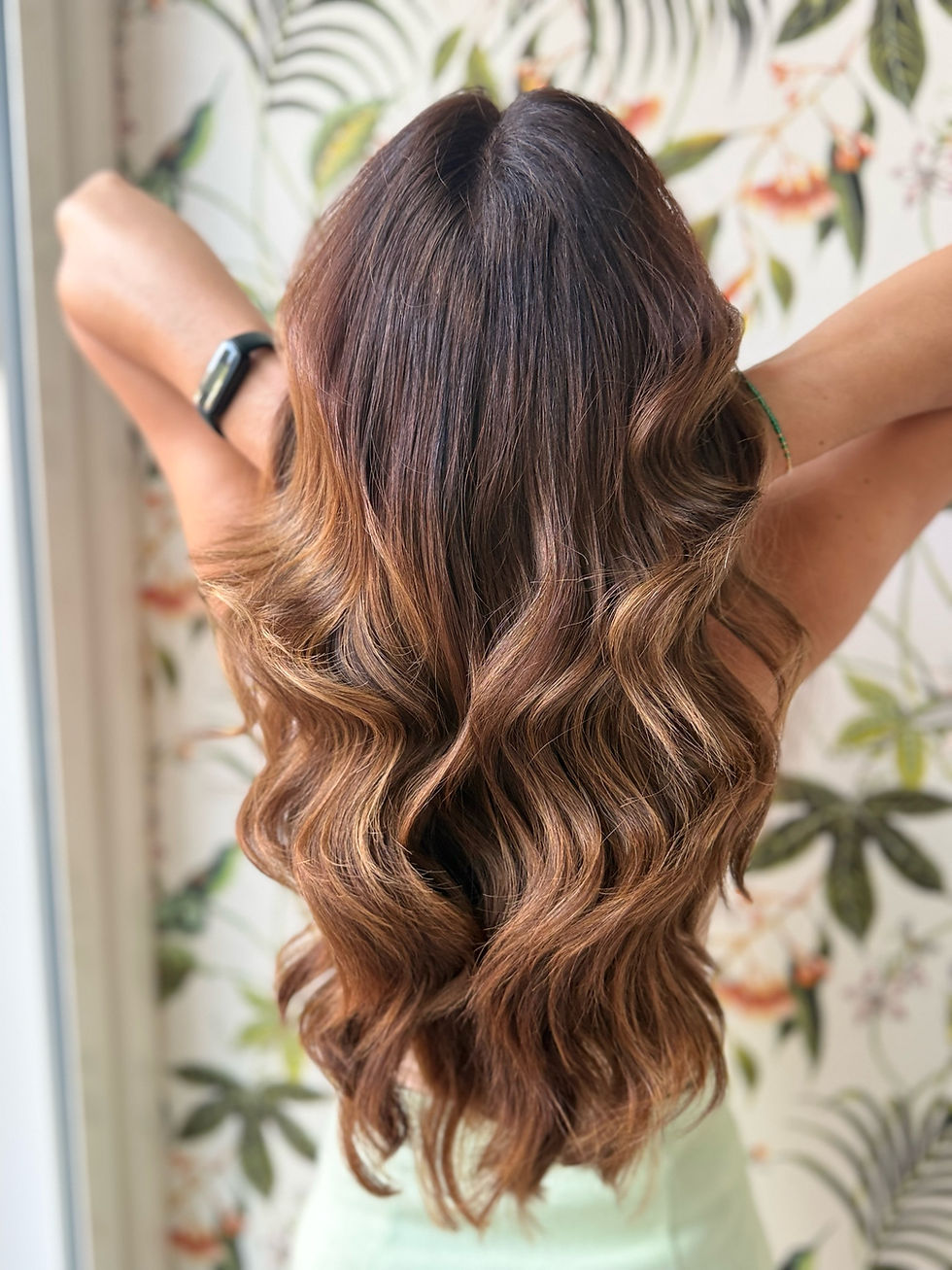 Woman seen from behind with long wavy brown hair and raised arms, in front of floral wallpaper.