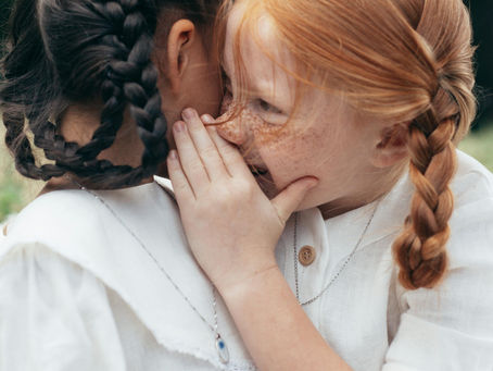 Two children with braided hair whisper and giggle outside. Both wear white shirts. The mood is playful and secretive.