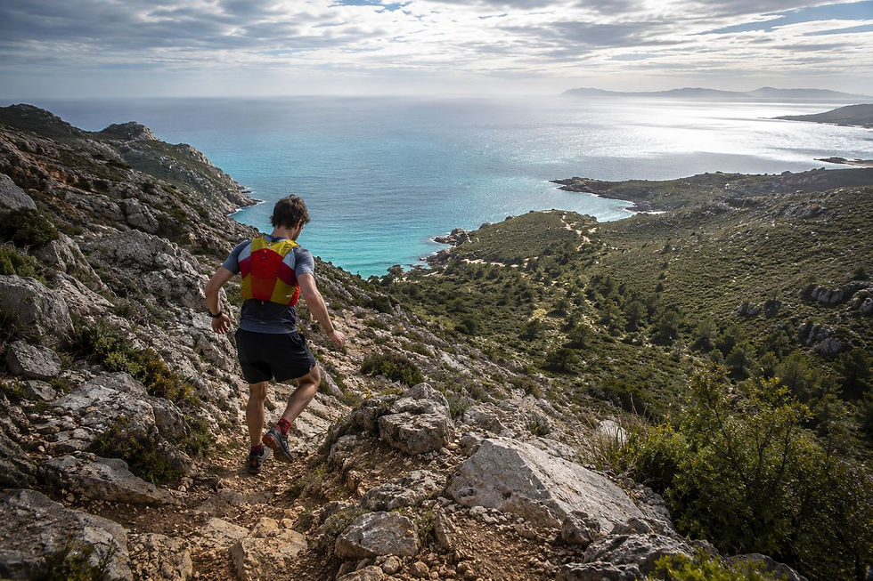 Trail runner in red-yellow vest descends rocky cliff path above turquoise sea and islands.
