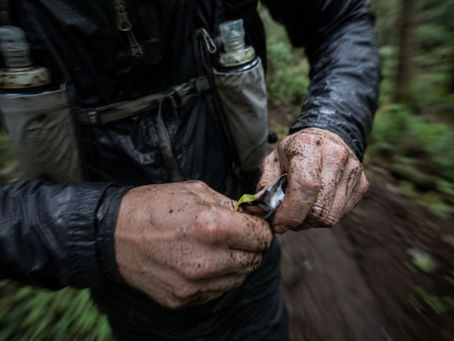 Muddy hands open a snack in a forest. Person wears a dark jacket with water bottles attached. The background is green and blurred.