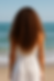 Woman with curly hair in a white dress stands facing the ocean. Blue sky and gentle waves create a serene beach setting.