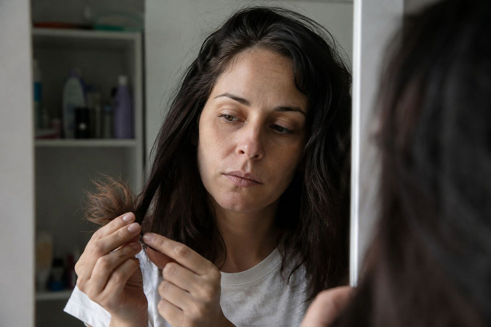 Woman in white shirt examines her hair in front of a bathroom mirror. She looks thoughtful. Shelves with toiletries are in the background.