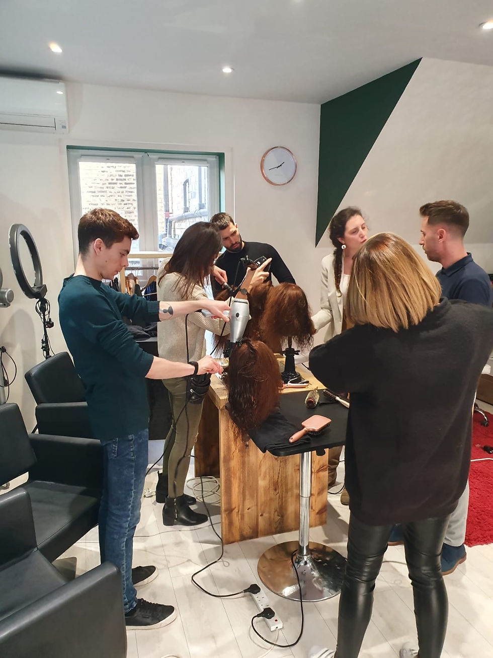 People styling a mannequin head with a blow dryer and tools in a salon. Light decor, clock on wall, and focused atmosphere.