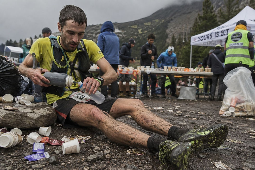 Runner in yellow shirt sits on muddy ground, refilling water bottle during a race. Volunteers and supply table in background. Overcast sky.