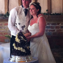 A bride and groom in formal attire stand smiling by a unique, dark-toned wedding cake.