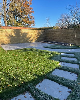 A sunlit garden with lush green grass and stepping stones leading to a wooden fence.