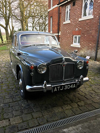 Classic black vintage car with chrome accents parked on a cobblestone driveway beside a red brick building