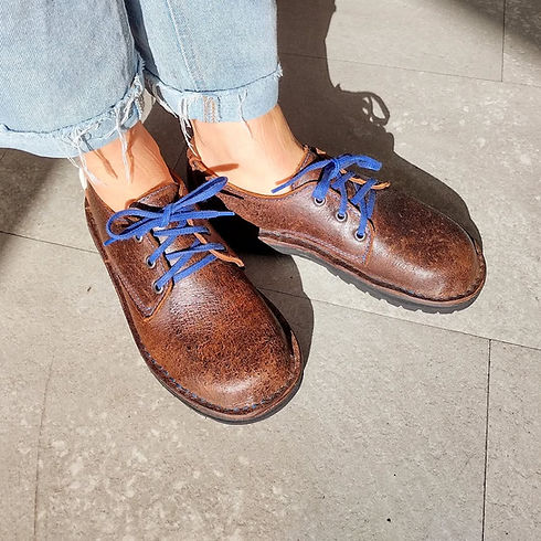 A person wears faded blue jeans and brown leather shoes with blue laces, standing on a gray tiled floor