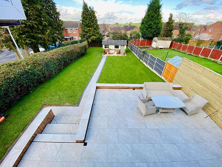 backyard with a large patio featuring wicker furniture. A lush green lawn and garden shed are enclosed by tall hedges and fences.