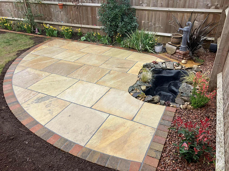Curved stone patio beside a small pond, with colorful plants and a water feature statue in a garden setting