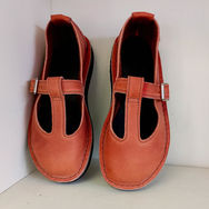 Two vibrant orange T-strap leather shoes with black soles are neatly placed side by side against a white background