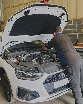 A mechanic leans over the open hood of a white Audi, inspecting the engine.