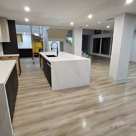 White quartz, dark cabinetry, full-height stone. A kitchen designed for everyday life — and for the moments worth Modern open kitchen with white quartz waterfall island, dark custom cabinetry, stone surround, and hardwood floors