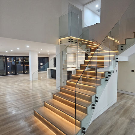Open-plan living area with LED-lit oak and glass staircase, wide-plank hardwood floors, and floor-to-ceiling windows