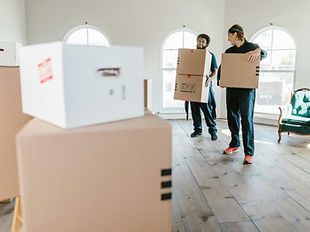 Two people carrying moving boxes in a bright room.