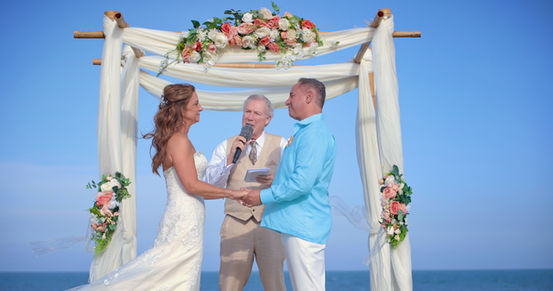 Bride and groom during their wedding ceremony in front of a four-post bamboo arbor with ivory sheers and coral accents on the beach in St. Croix, USVI, with the groom wearing aqua.