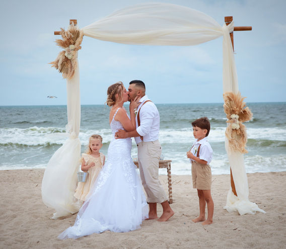 Bride and groom sharing their first kiss under a bamboo wedding arbor with their children on the beach in St. Croix, USVI during a destination beach wedding ceremony.