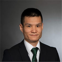 Headshot of Yugo Iwamoto, with short dark hair, wearing dark suit, white dress shirt, and green tie, looking at the camera with a slight smile against natural grey background.
