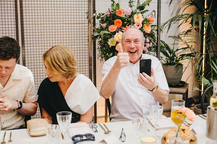 Groom’s father giving a thumbs up and holding a smartphone while the groom’s mother looks at her son’s hands. The Hackney, London.