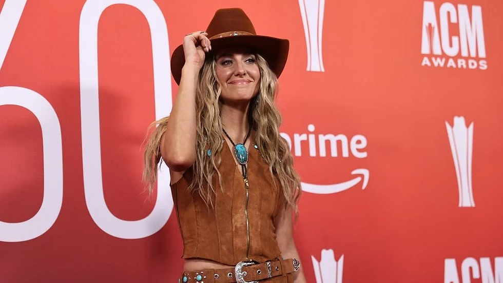 Lainey Wilson in a brown cowgirl outfit smiles, adjusting her hat on a red carpet. "ACM Awards" and "Prime" logos visible in the background.