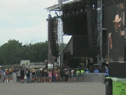 XRoads41 concert with a crowd in front of a stage showing a performer on a large screen. Green trees and a cloudy sky in the background.