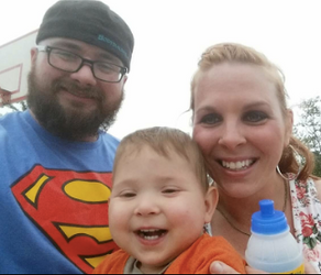 Smiling family outdoors; man in Superman shirt, woman, and child with a blue-topped bottle. Overcast sky and basketball hoop in background.