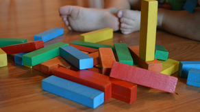 Colorful wooden blocks scattered on a wooden floor near a child’s bare feet. The scene is playful and casual, with warm tones.