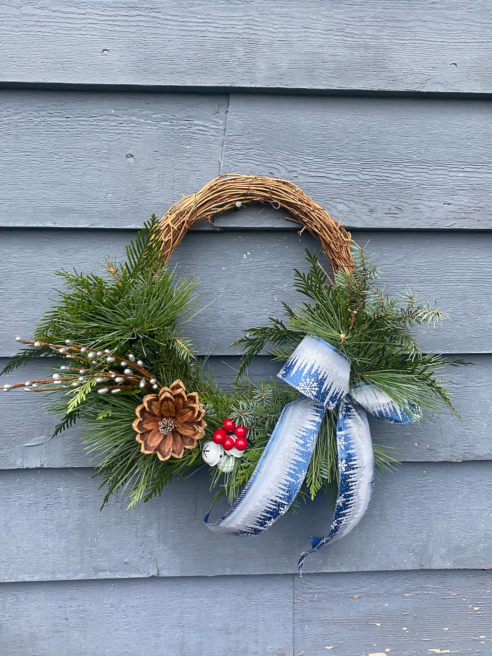 Rustic wreath with green pine, pinecone, blue bow on blue siding.