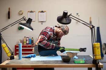 High-fidelity wide shot of an artisan working on a guitar neck at a specialized workbench with dual task lamps.