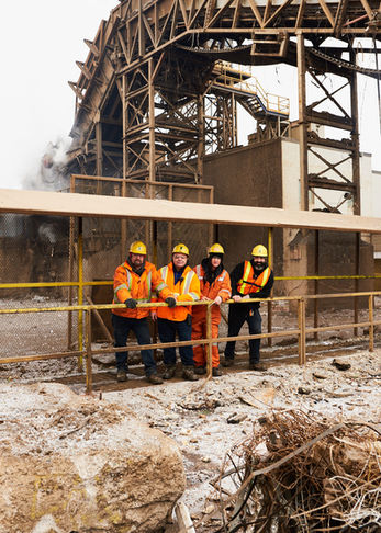 Group environmental portrait of four industrial staff members standing behind a safety railing at an outdoor metal processing site.