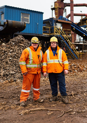 Candid environmental portrait of two industrial workers in orange high-visibility gear standing before heavy machinery at MMM Metal.