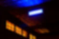 The image depicts a dimly lit interior, with glowing windows casting soft light. The focal point is a  blue, rectangular sign mounted on the ceiling. The sign features the lettering '+ΕΡΓΕΙΟ,' clearly identifying the location as the Synergeio Performing Arts Centre. 