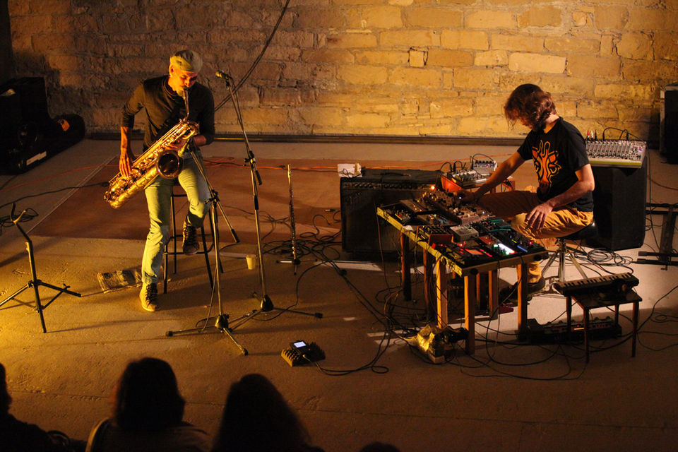 Two male musicians perform on a lit stage with a concrete floor, one playing the saxophone and the other creating electronic music, set against a stone wall backdrop.