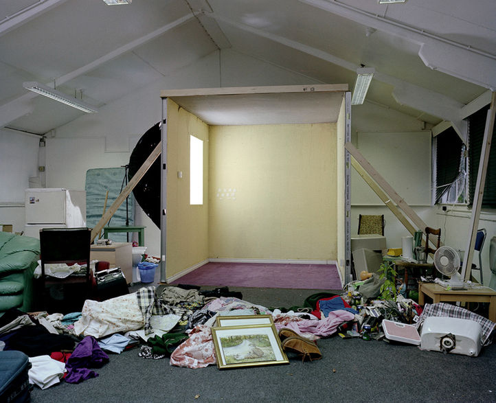 Partially dismantled installation showing an empty constructed room beside scattered objects, clothing, and framed images on the studio floor.