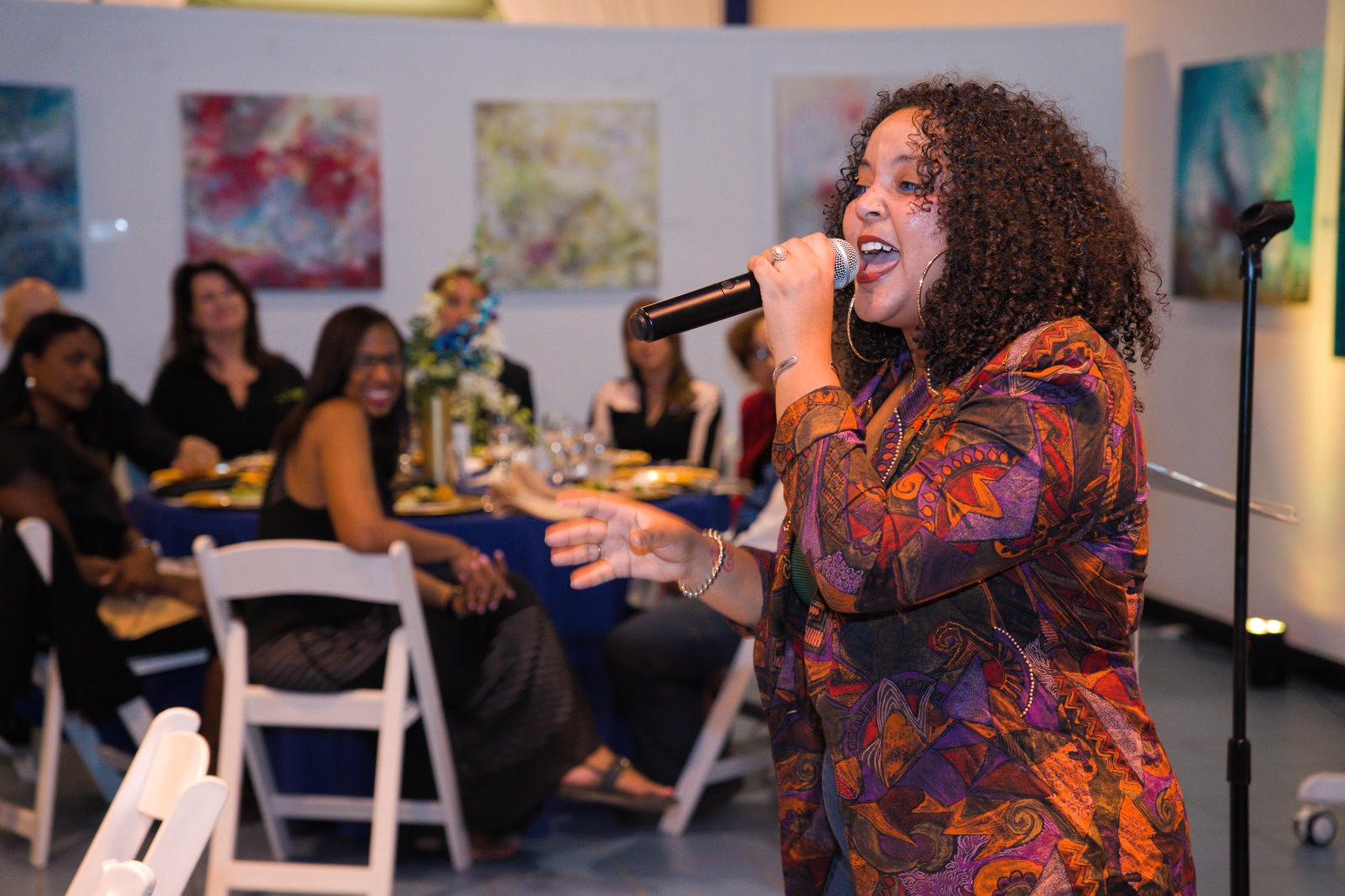 Frankie Red performing in a vibrant patterned jacket. Seated guests in the background watch, with colorful art on the walls.