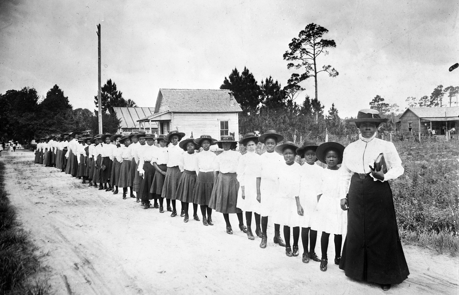 A historic photograph showing a line of young Black girls standing in uniform outside a small school building, guided by their educator, reflecting discipline, dignity, and the early foundations of a school built through love, faith, and determination.