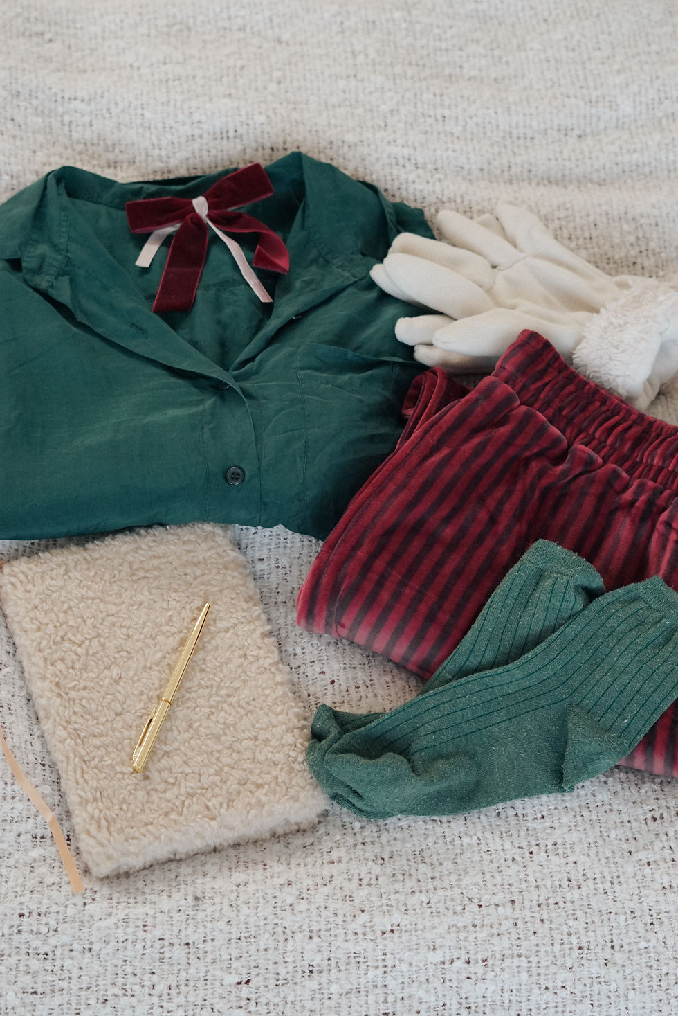 Green shirt with red bow, cream gloves, red striped pants, and green socks on a textured surface. A gold pen rests on a fuzzy notebook. Cozy vibe.