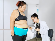 A doctor measures a woman's waist with a tape in a bright, clinical room. The woman stands while the doctor sits with a stethoscope around her neck.
