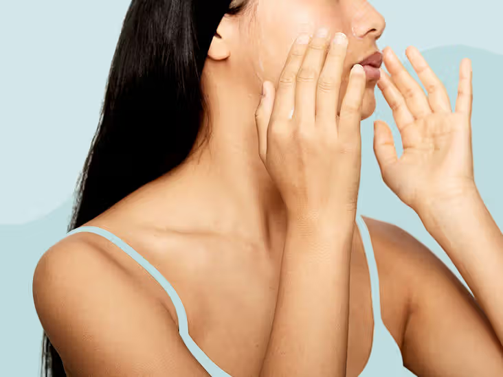 Woman applying skincare product to her face with both hands. She has long dark hair and wears a light blue strap. Soft blue background.